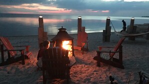 A beach scene with a fire pit, wooden chairs, and a hammock.