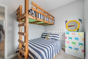 Ein Stockbett mit Holzleiter, einer gestreiften Bettdecke und einer weißen Kommode mit bunten Palmenmotiven.