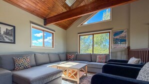 A cozy living room with a grey sectional sofa, a wooden coffee table, and a dark armchair. There are two windows providing natural light and a view of trees. - Panoramic views, Hot Tub, Sauna, Firepit, Sand Vball, Foosball, Dunes Nat`l Park (Miller Beach)