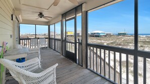 A covered porch with wicker furniture and a table, overlooking a beach with buildings and a clear sky.