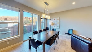 A modern dining area with a large table, chairs, and a bench. There are large windows providing a view of the outdoors.