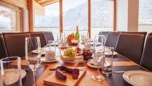 A dining table set with plates, glasses, and a variety of food items, with a mountain view through large windows. - Chalet Nepomuk (Zermatt)