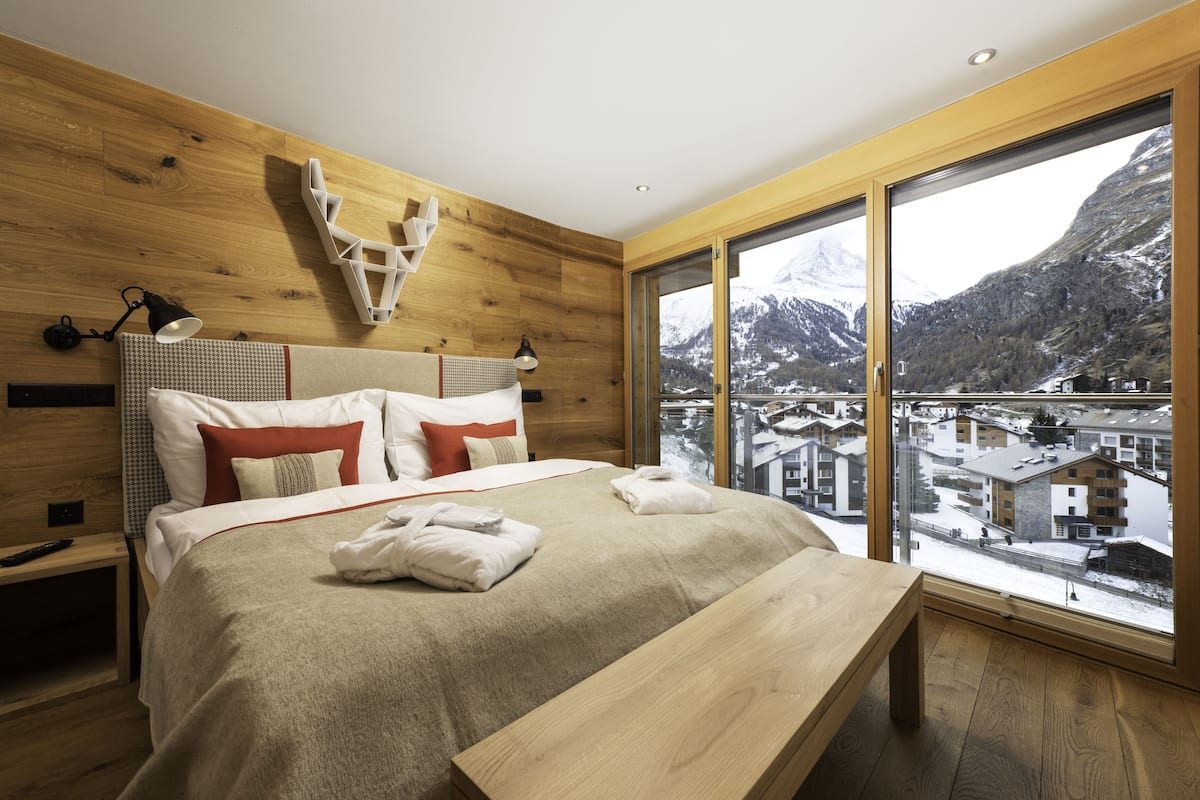 A modern bedroom with a wooden headboard, a large bed with multiple pillows, a bedside table, and a view of a snowy mountain town through large windows.
