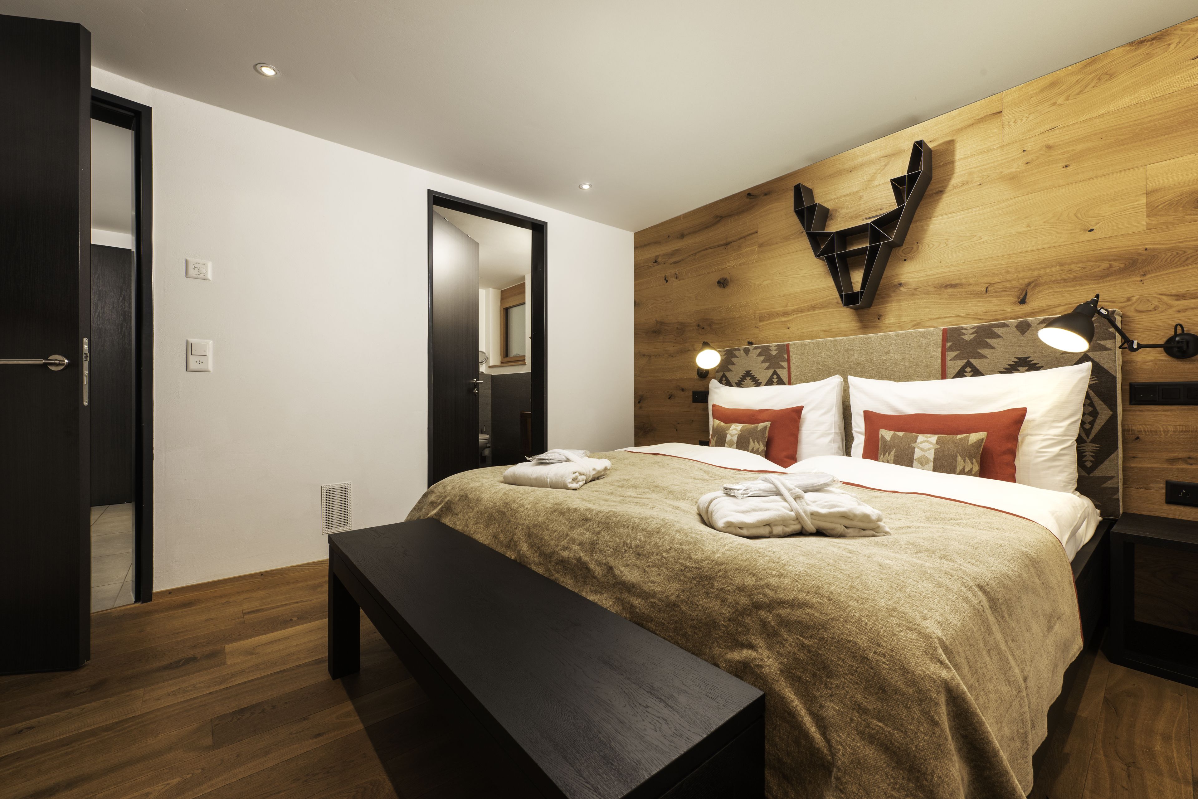 Quarto com cabeceira de madeira, cama com almofadas brancas e vermelhas, um banquinho e uma mesa de cabeceira com abajur.