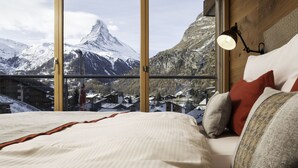 A bedroom with a large window offering a mountain view, a bed with pillows, and a wall-mounted reading lamp. - Chalet Nepomuk (Zermatt)