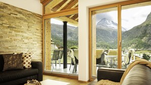 A modern living room with a black leather sofa, a white coffee table, and a view of a mountainous landscape through large glass doors. - Chalet Nepomuk (Zermatt)