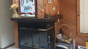 Un interior de madera con una chimenea, un cartel enmarcado y un jarrón con flores secas.