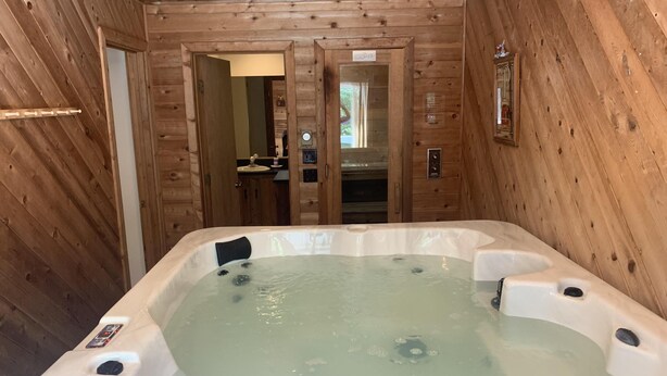 Interior de una cabaña de madera con jacuzzi, paredes de madera y un baño visible con lavamanos y espejo.