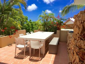 An outdoor patio with a long table and chairs, surrounded by lush greenery and a stone wall.