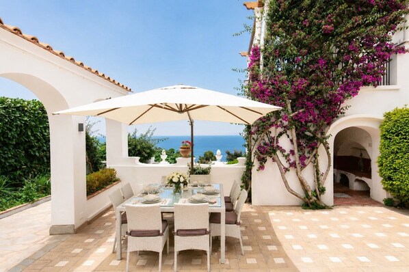 A patio with a table set for four, surrounded by white columns and arches, overlooking the sea.