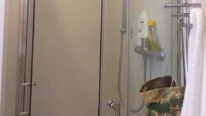 Cabina de ducha con paredes de vidrio, un cabezal de ducha de mano, un grifo empotrado en la pared y un perchero con una toalla blanca.