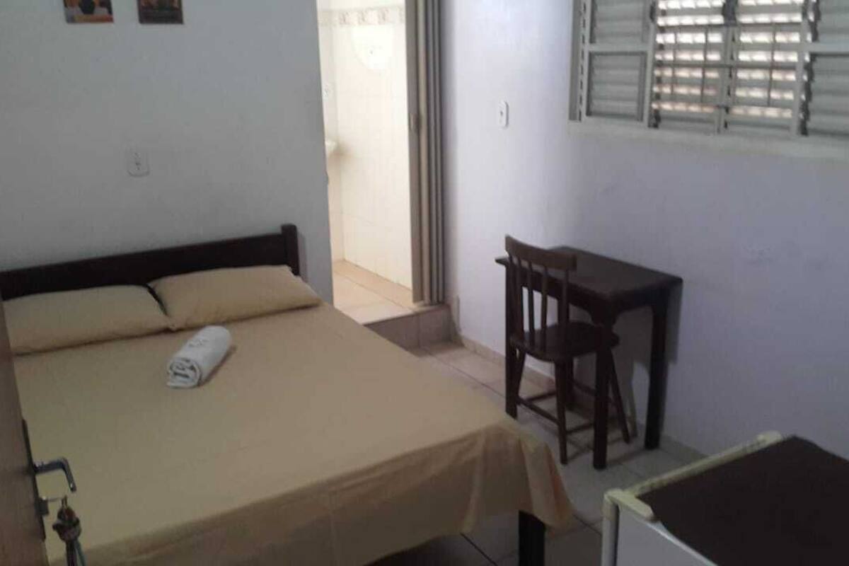 Une chambre avec un lit, un bureau et une chaise. Il y a une fenêtre avec des stores, une porte et une petite table. Une serviette est posée sur le lit.