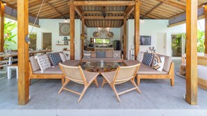 Un’area salotto esterna coperta con un tavolo in vetro, sedie di legno e un divano con cuscini decorati.