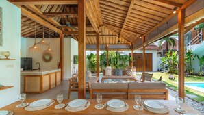 Un'area pranzo all'aperto coperta con tavolo in legno apparecchiato per sei persone, un divano e una piscina sullo sfondo.