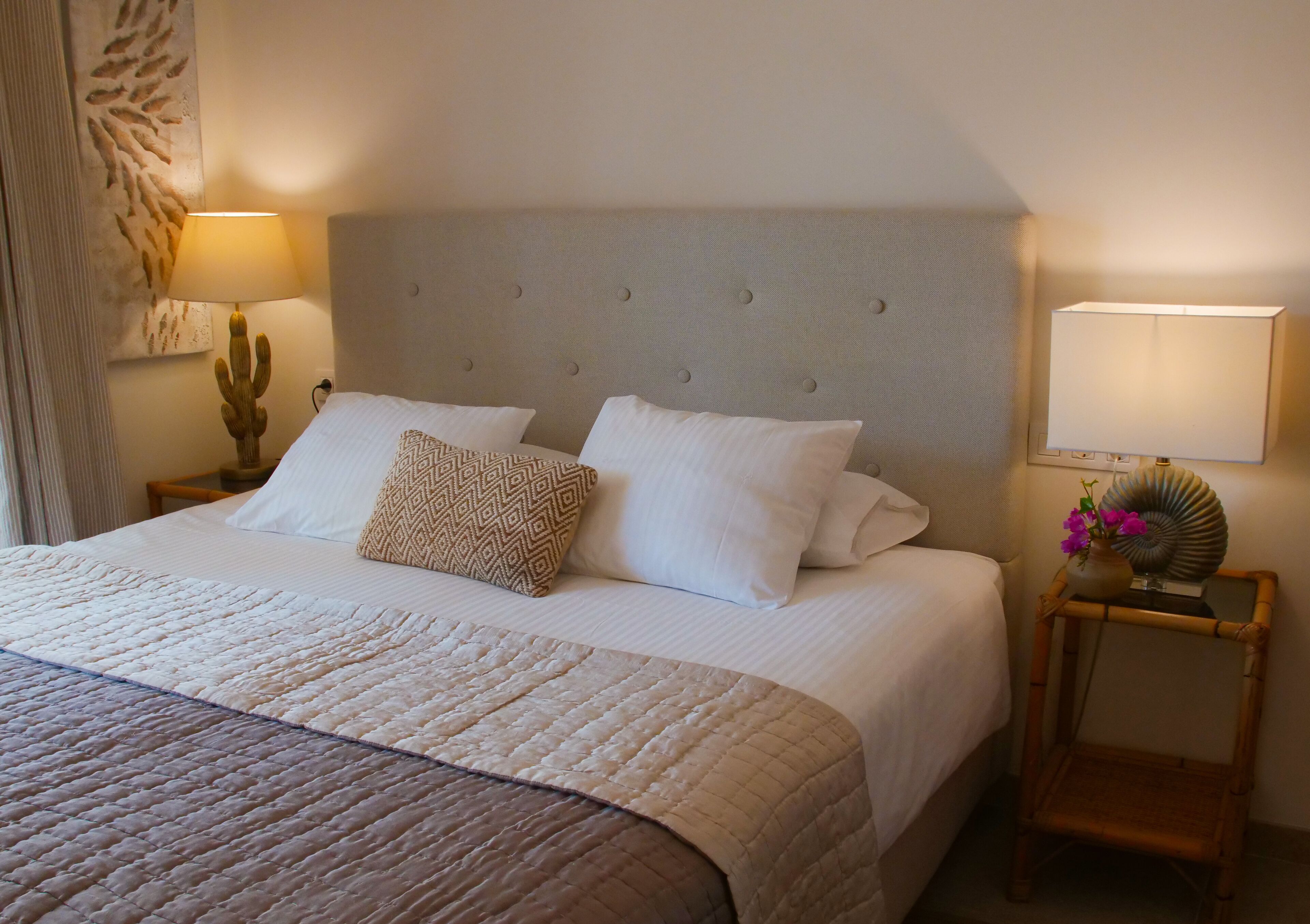 A neatly made bed with a quilted bedspread, two pillows, and a decorative pillow. There is a bedside lamp on the left and a small table with a vase of flowers on the right.