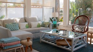 A well-lit living room with a sofa, a glass coffee table, a wicker chair, and a small table with a lamp.