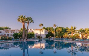 Zona de piscina con palmeras y sillas de descanso, rodeada de edificios de arquitectura mediterránea.
