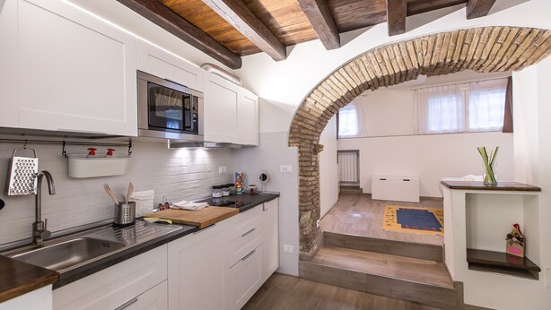 Een moderne keuken met witte kastjes, een roestvrijstalen spoelbak en een magnetron. Een stenen boog leidt naar een eethoek met een houten tafel en stoelen.