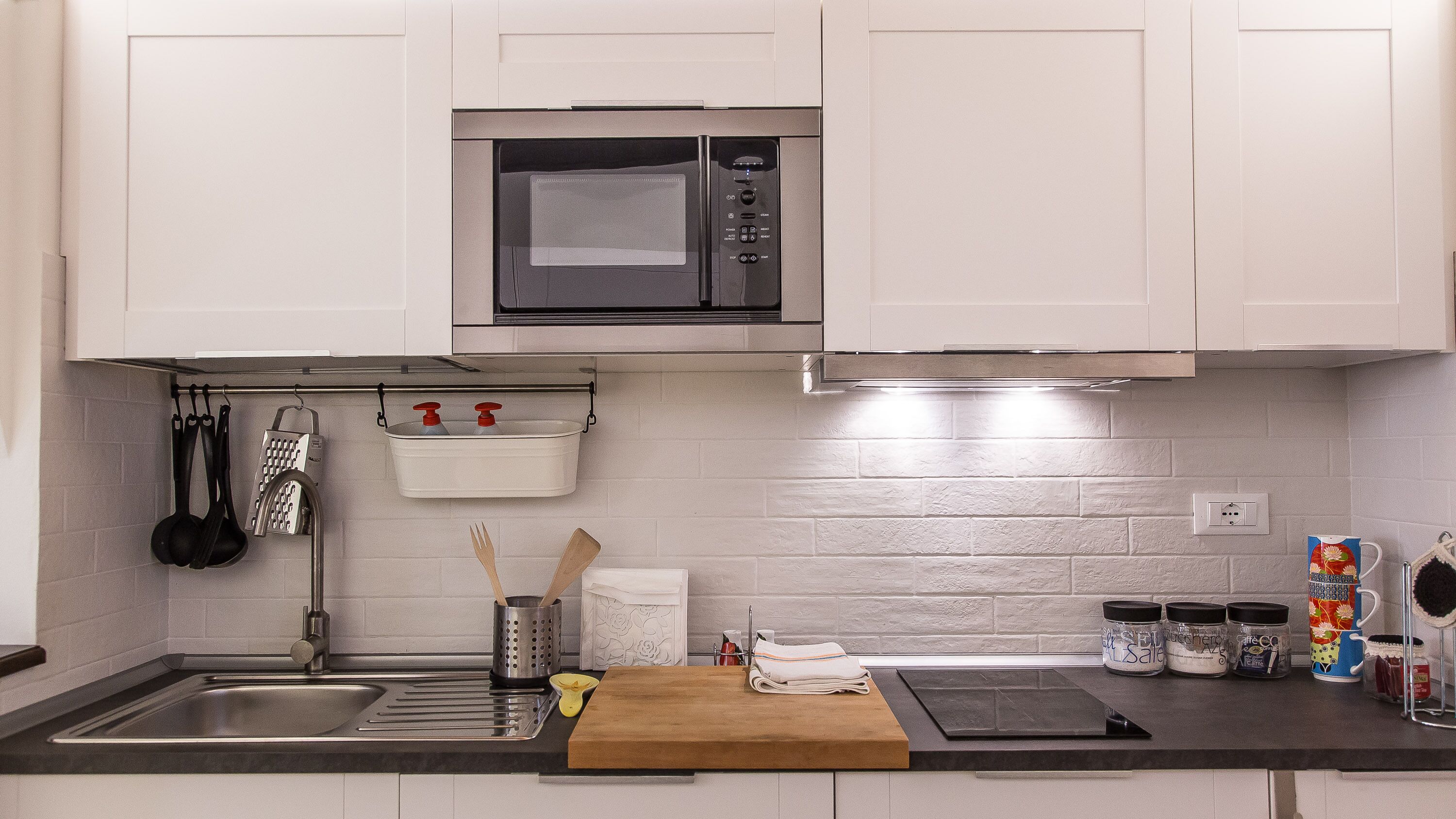 Una cocina moderna con armarios blancos, un microondas de acero inoxidable, un lavamanos y diversos utensilios de cocina.