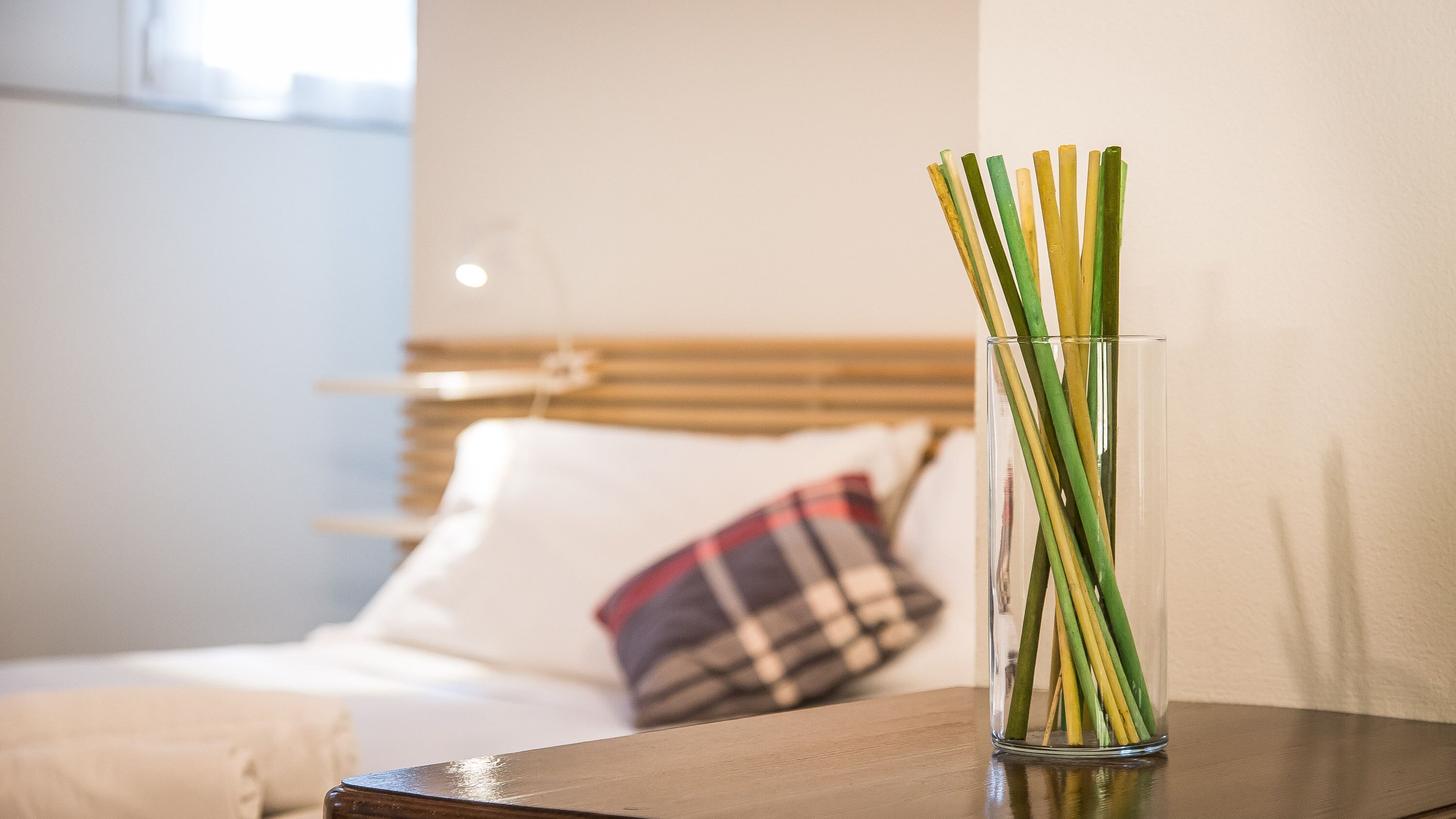 A clear glass vase with green and yellow stems on a wooden table in a hotel room.