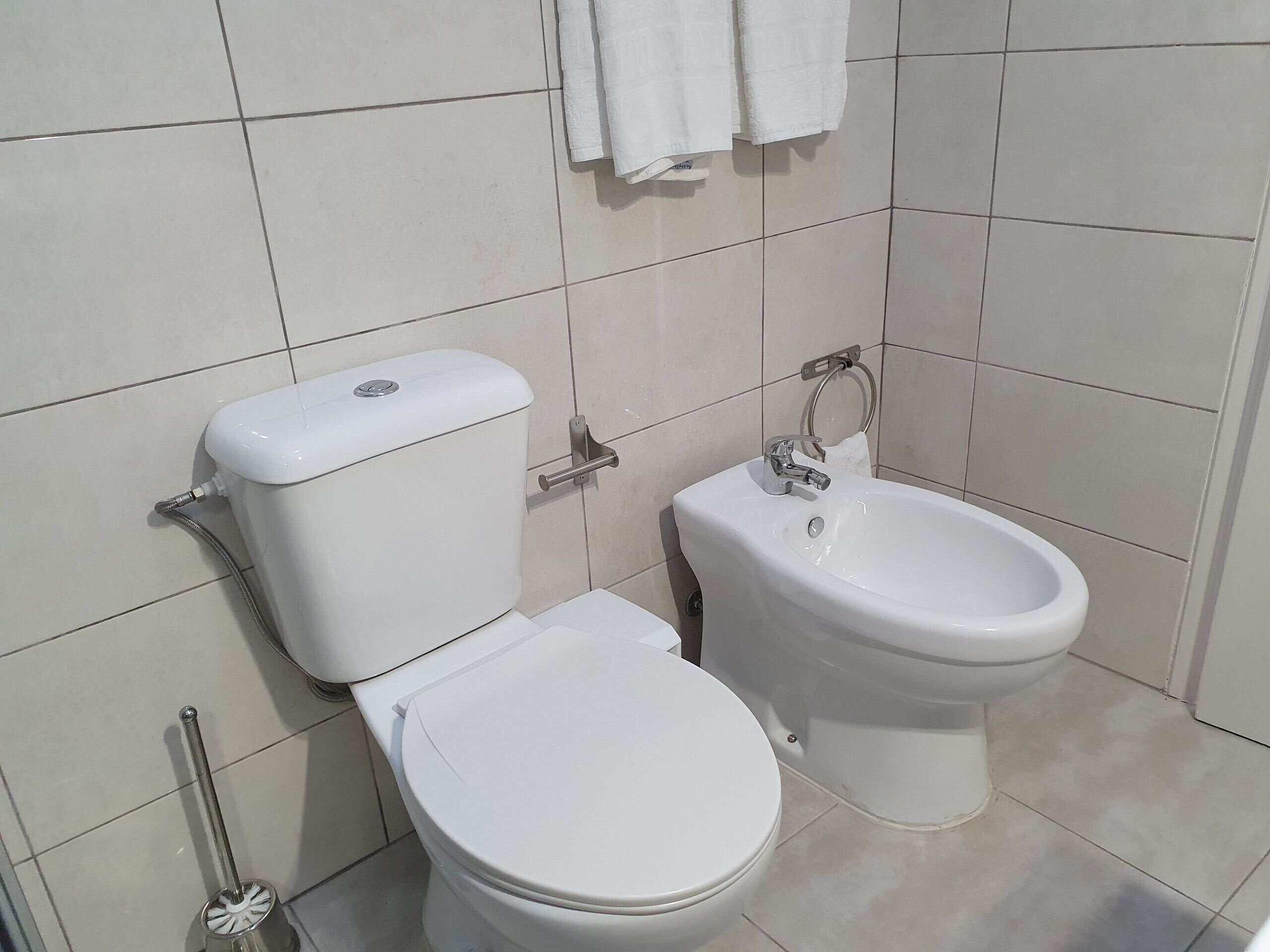 A bathroom with a toilet and bidet, both white, and a toilet brush.
