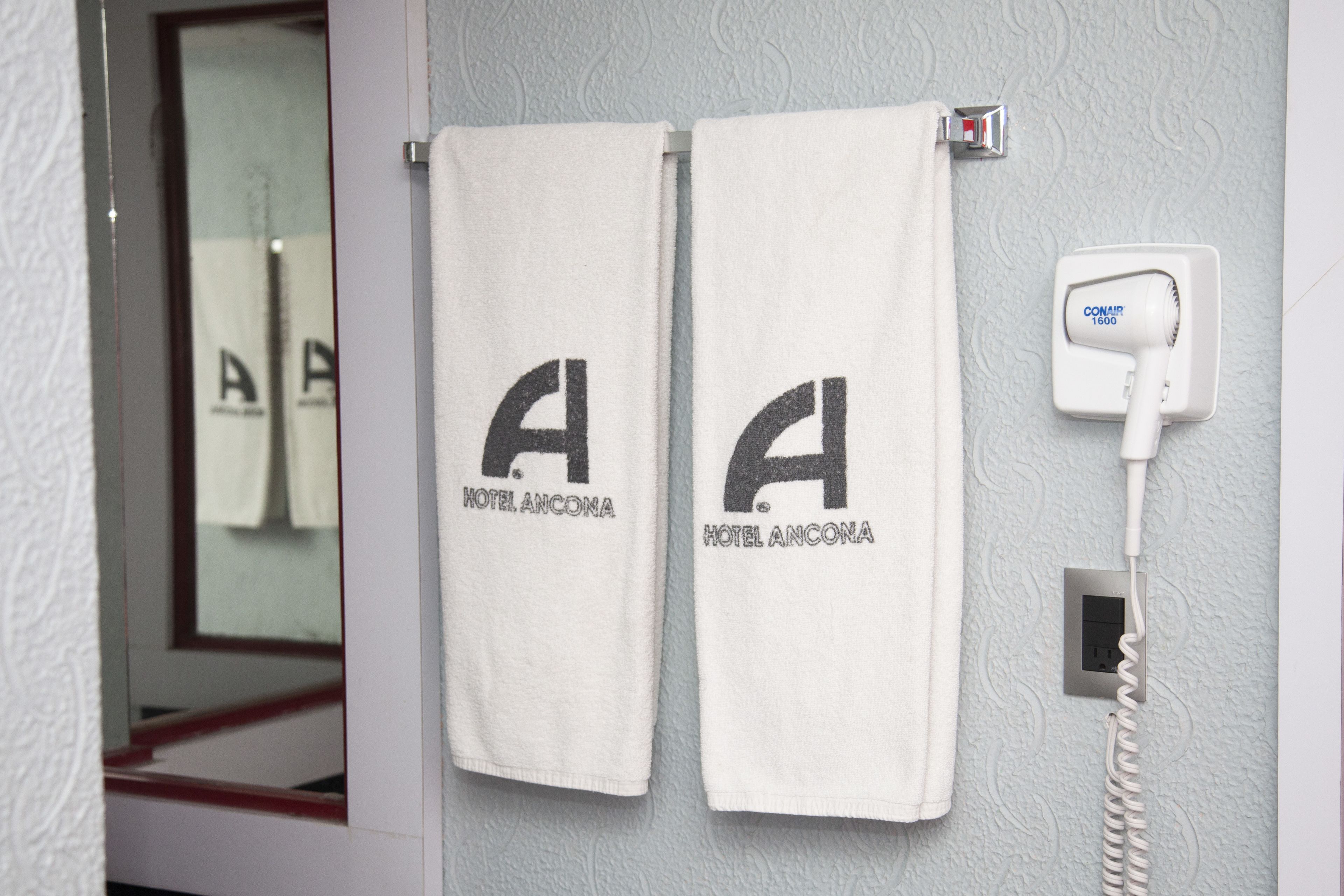 Duas toalhas brancas penduradas na parede com o logotipo do Hotel Ancona, um secador de cabelo e uma tomada.