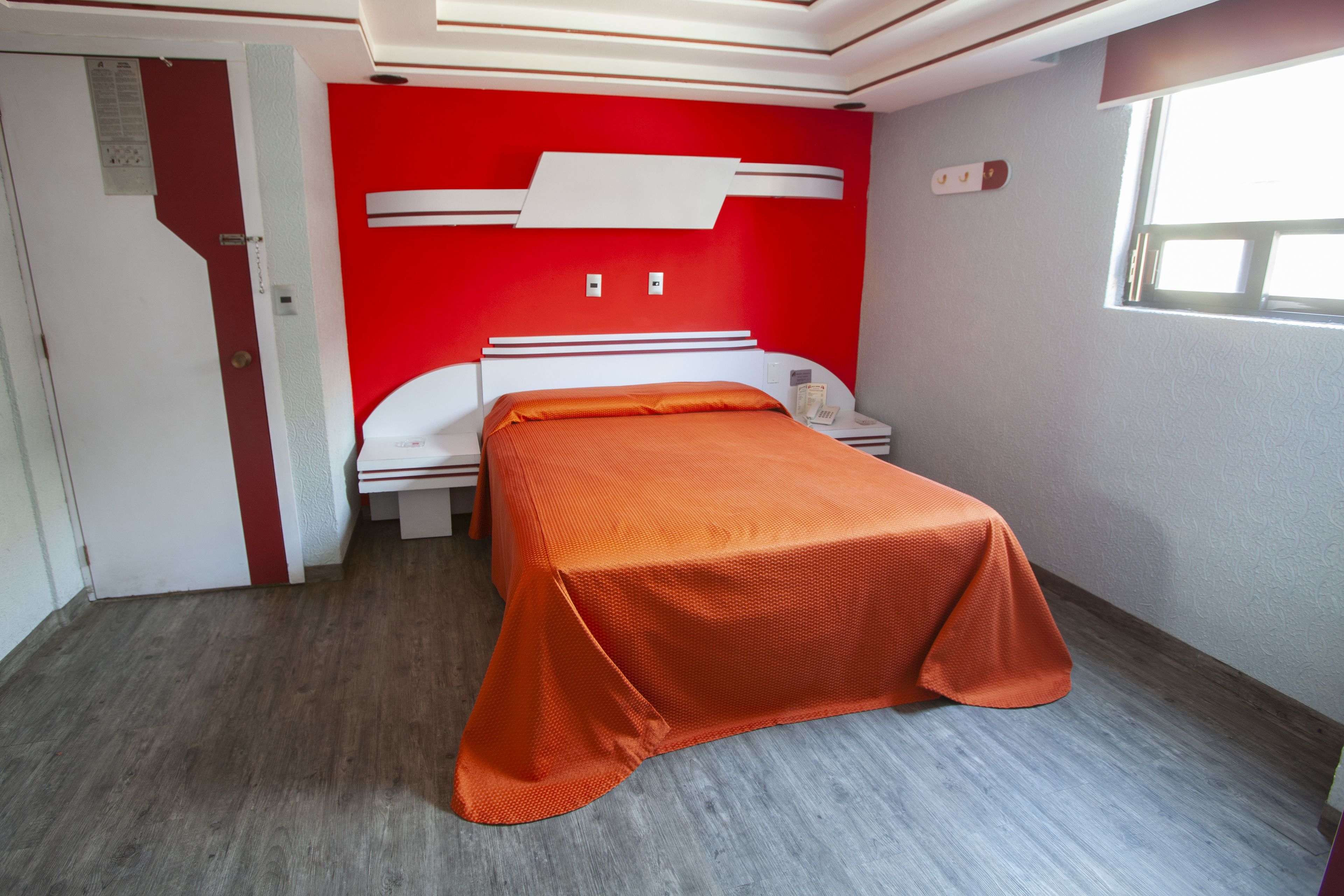 Una habitación con cama, mesita de noche, pared roja y ventana.