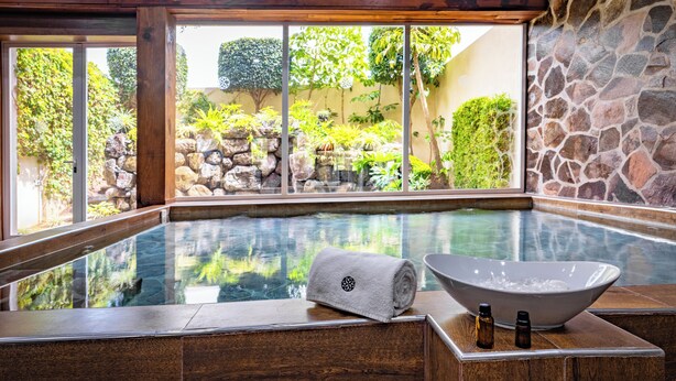 Een wellnessruimte met een bubbelbad, houten vloeren, stenen muren en uitzicht op een tuin met rotsen en beplanting.