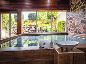 Un espace détente avec un jacuzzi, un plancher en bois, des murs en pierre et une vue sur un jardin agrémenté de rochers et de plantes.