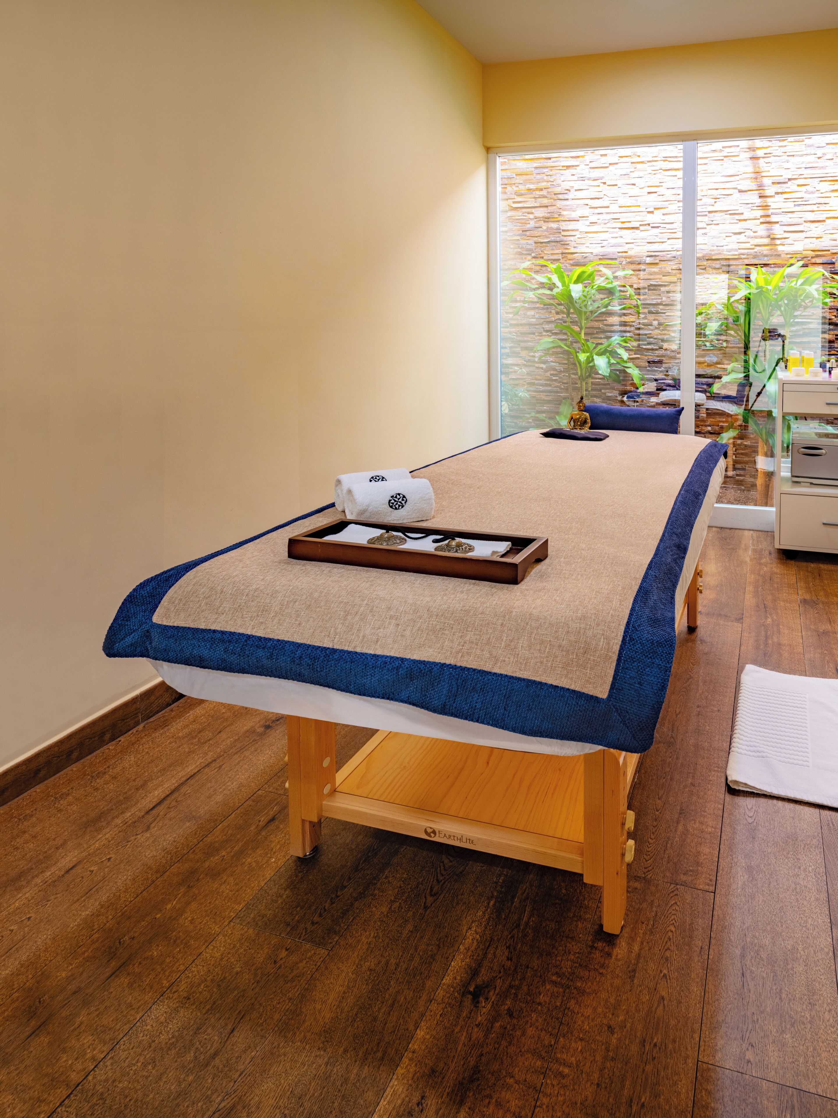 A massage table with a towel and a tray of massage tools in a room with wooden flooring and a view of plants through a glass wall.