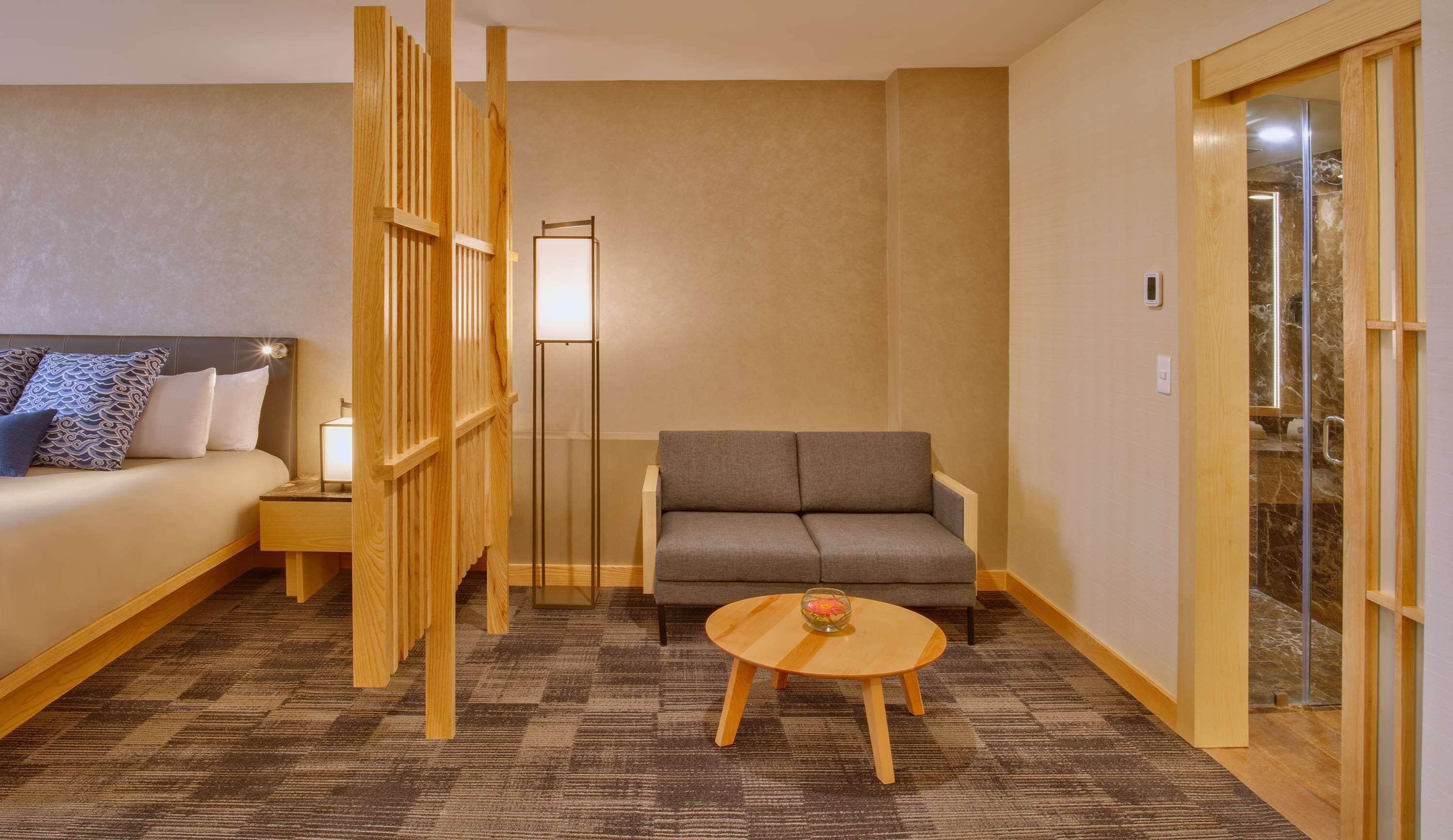 Habitación de hotel con cama, sofá, una mesita con un cuenco, lámpara de pie y una puerta corredera que da a un baño.