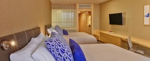 A hotel room with two beds, a desk, a TV, and a window with blinds.