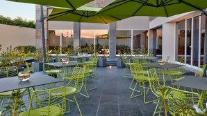 Een terras buiten met groene metalen stoelen en tafels, grote parasols en een uitzicht op de stadsachtergrond.