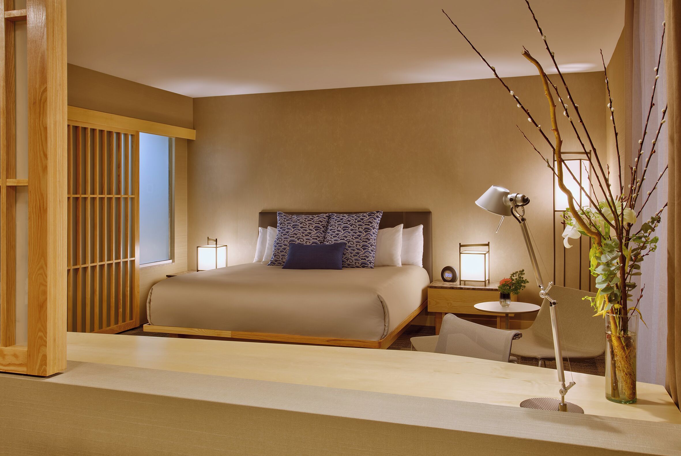 Une chambre d’hôtel bien aménagée, avec un grand lit, des lampes de chevet, une petite table et un vase décoré de branches.