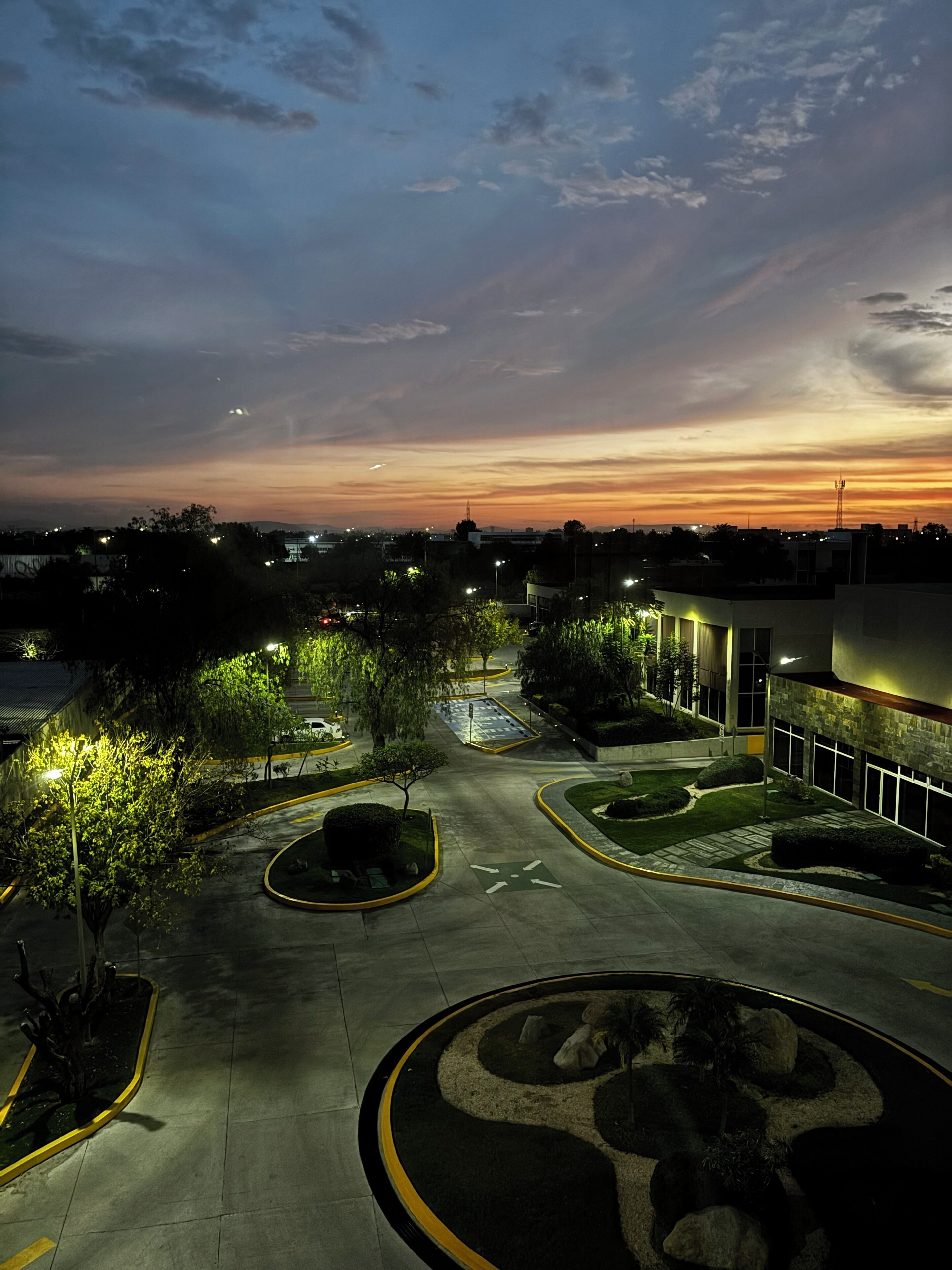 A parking lot with a landscaped area, a building with a modern design, and a sunset sky.