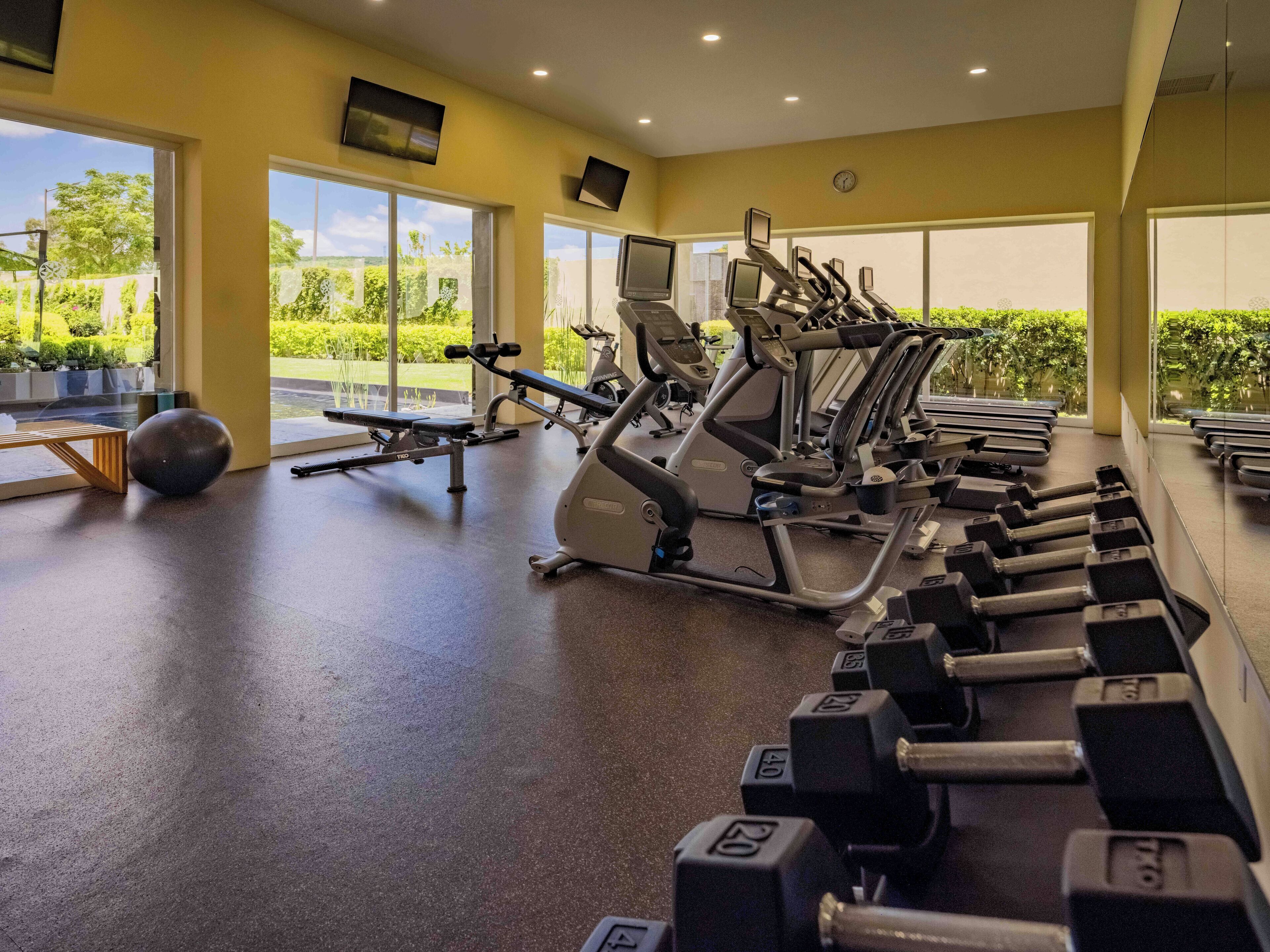 Un gimnasio con equipo de ejercicio, ventanas amplias y vistas a zonas verdes.