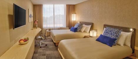 A hotel room with two beds, a desk with a flat-screen TV, a small table with a bowl of fruit, and a view of the city through large windows.