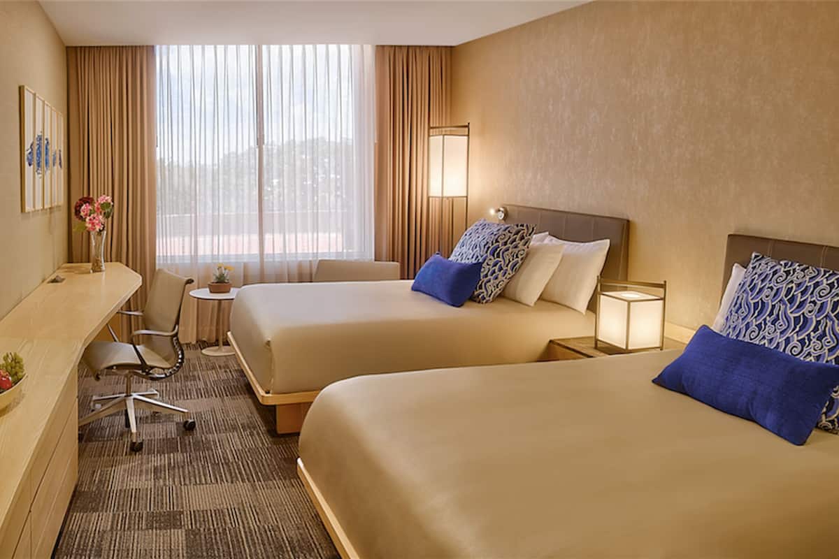 Une chambre d’hôtel avec deux lits, un bureau agrémenté d’un panier de fruits, une télévision et une fenêtre avec des rideaux.
