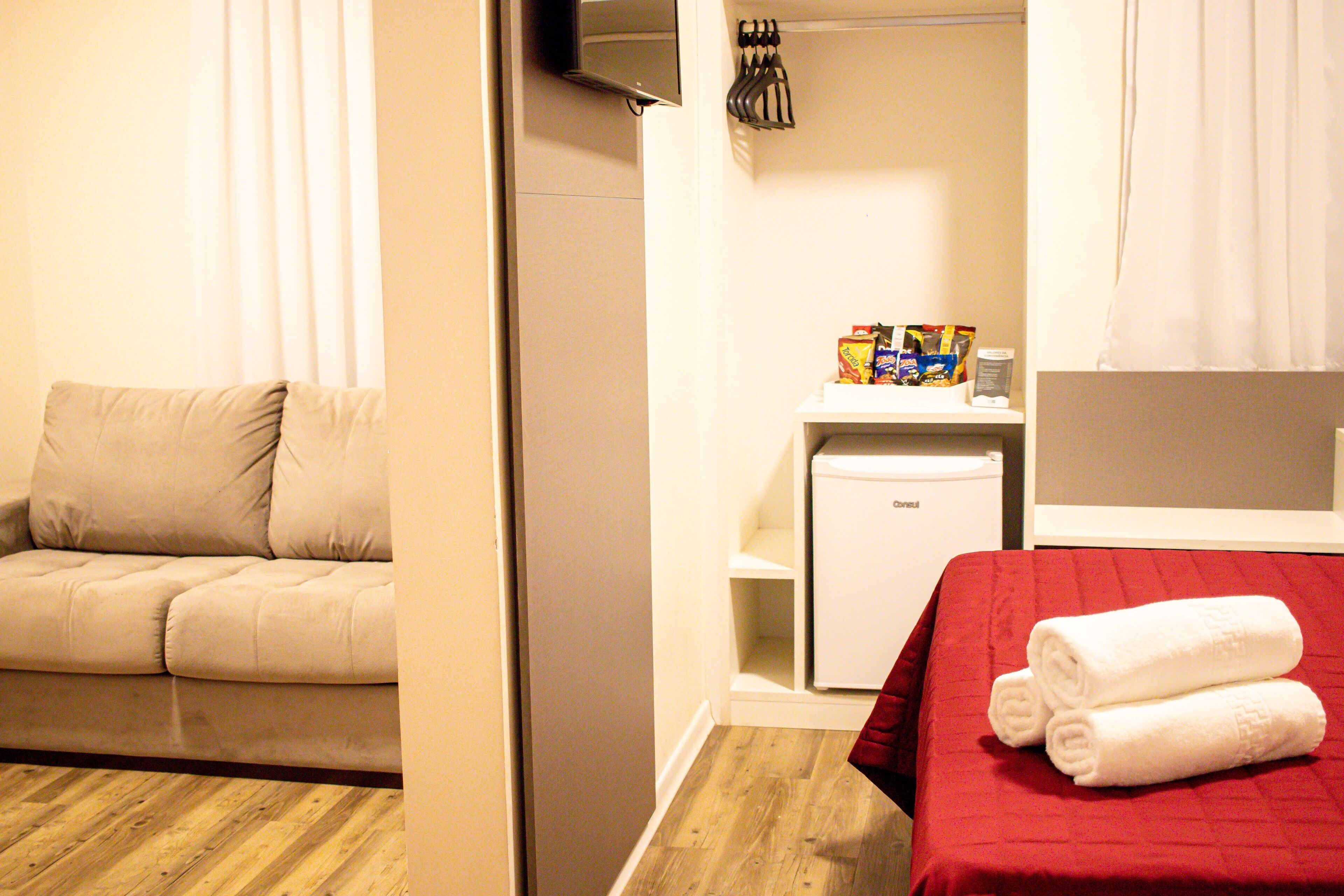 A hotel room with a wooden floor, a beige sofa, a small white refrigerator, and a bed with a red blanket and two rolled towels.