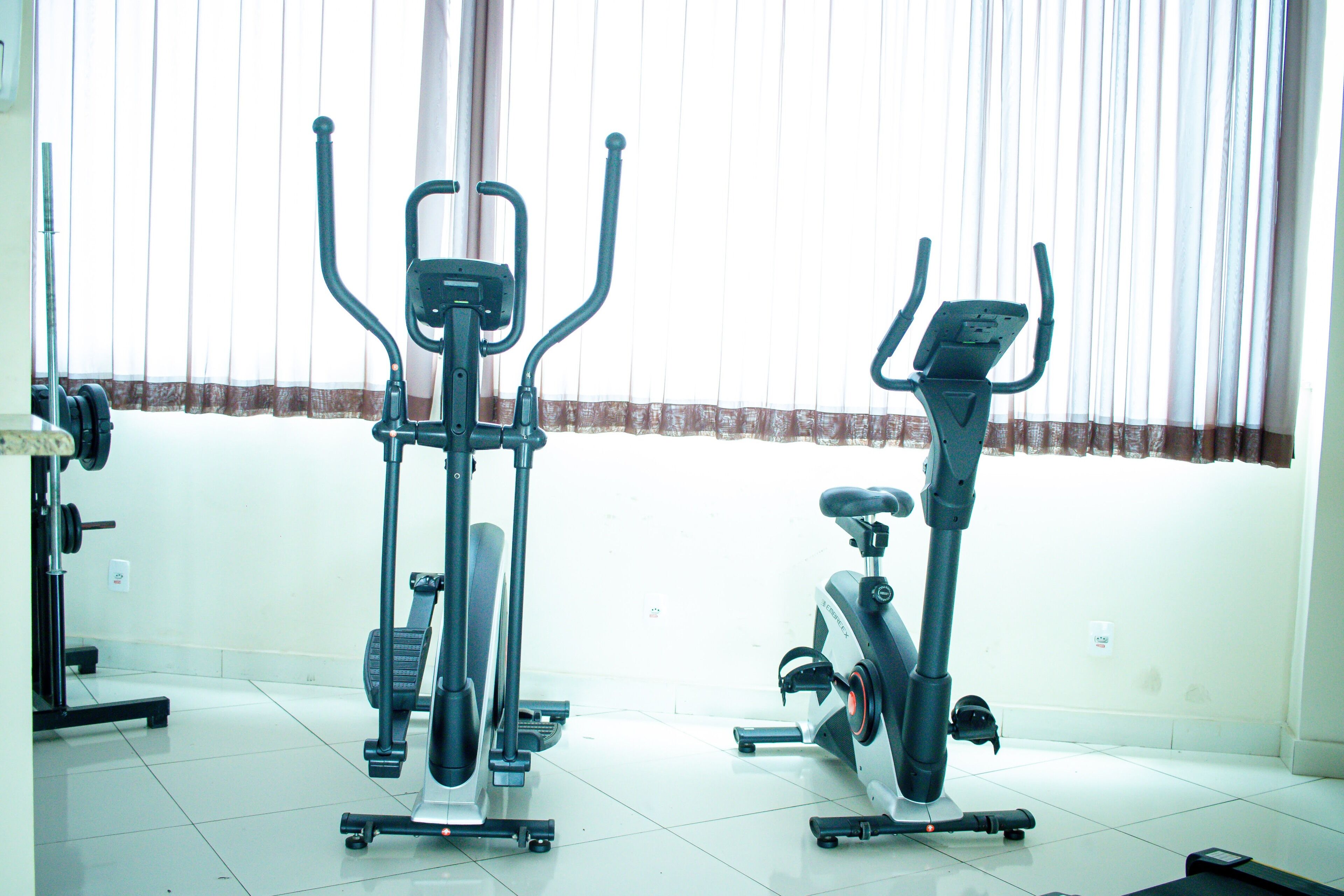 Two stationary bikes in a room with a tiled floor and a window with sheer curtains.