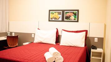 Una habitación de hotel con una colcha roja, dos almohadas blancas, una silla de madera, un escritorio con teléfono y dos cuadros en la pared.