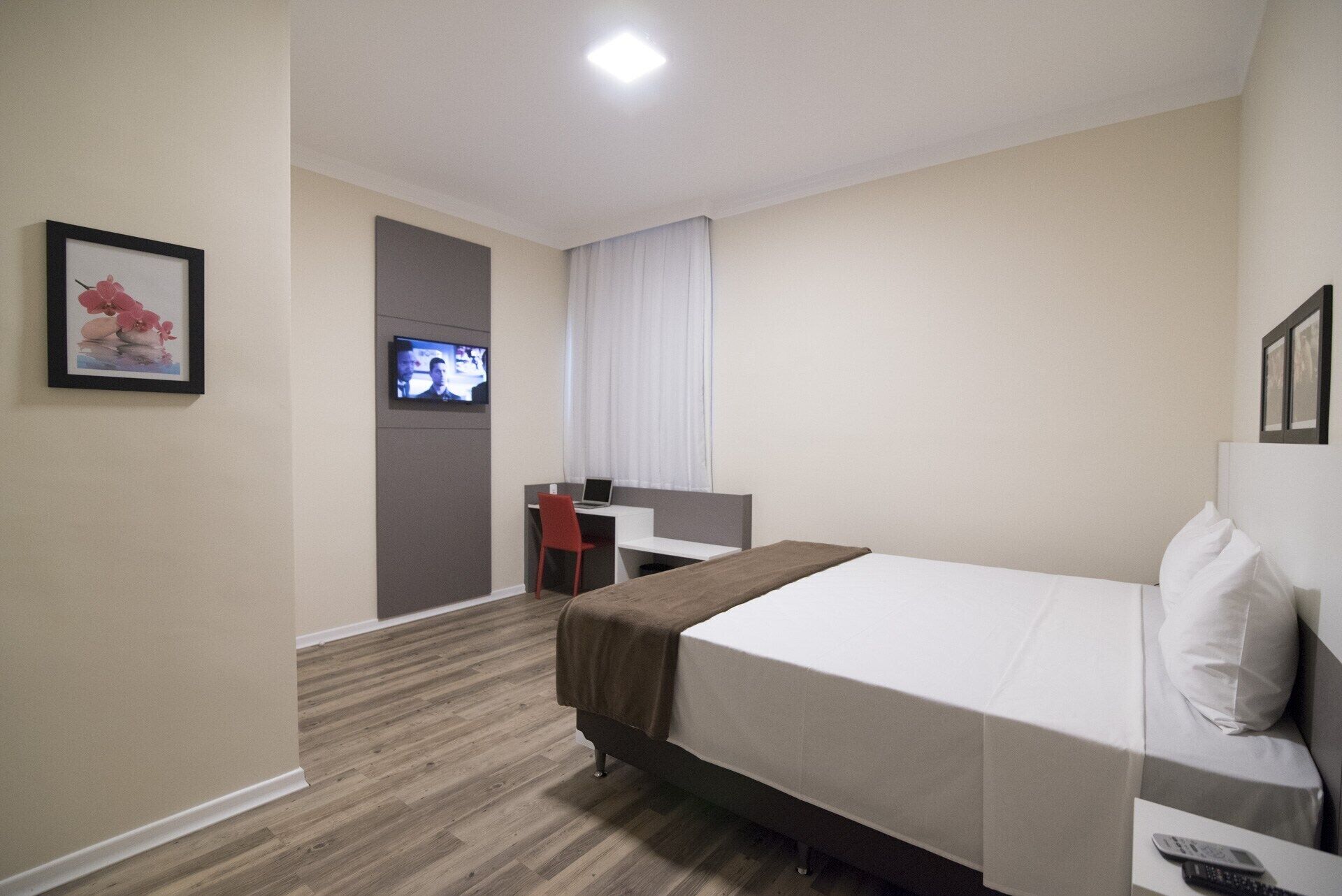 Habitación de hotel con cama, escritorio con silla, televisor montado en la pared y un cuadro enmarcado.
