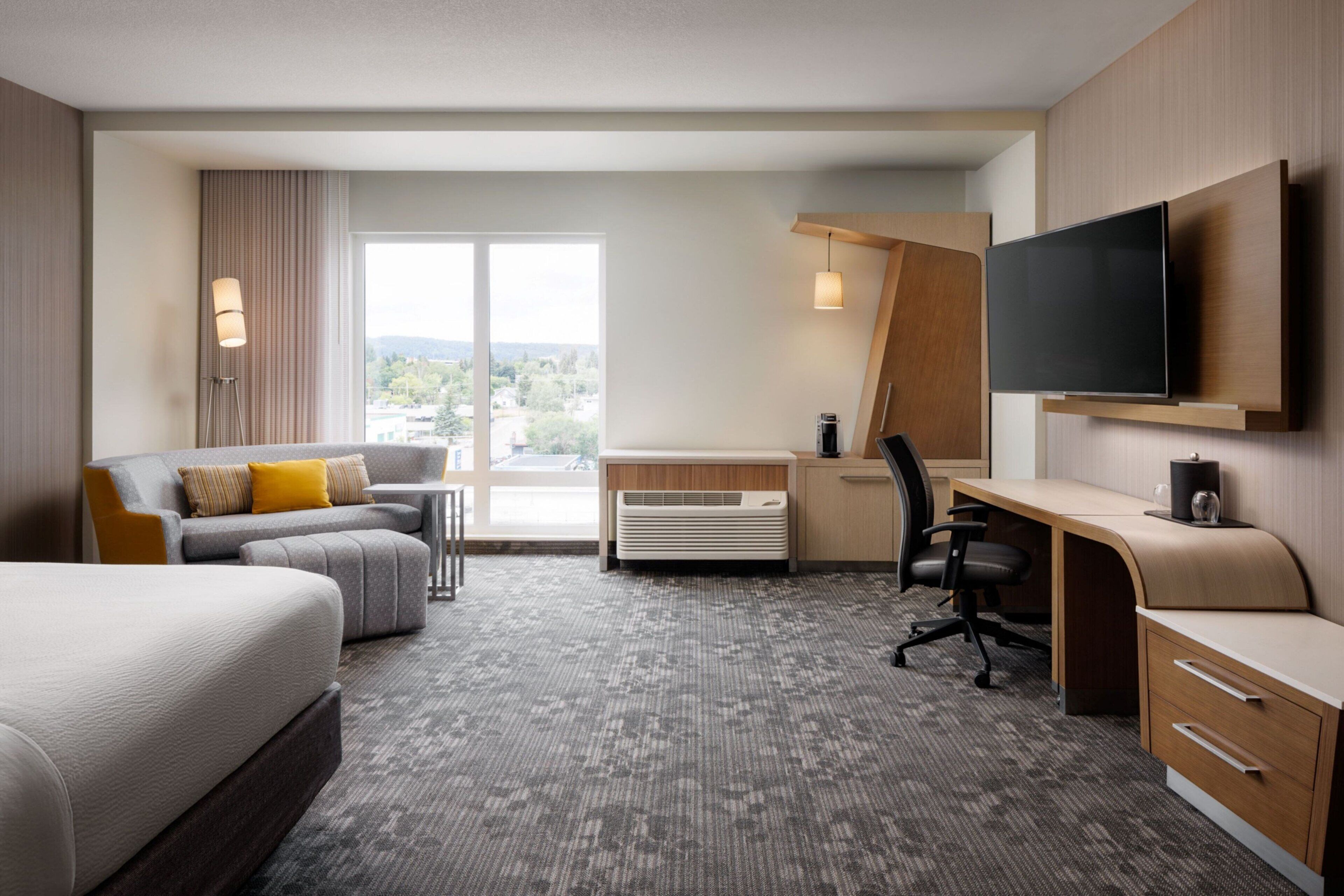 Habitación de hotel con cama, escritorio con silla, sofá, televisor montado en la pared y un amplio ventanal con vistas al exterior.
