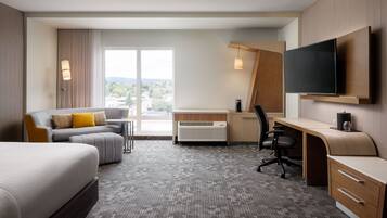 Habitación de hotel con cama, escritorio con silla, sofá, televisor montado en la pared y un amplio ventanal con vistas al exterior.