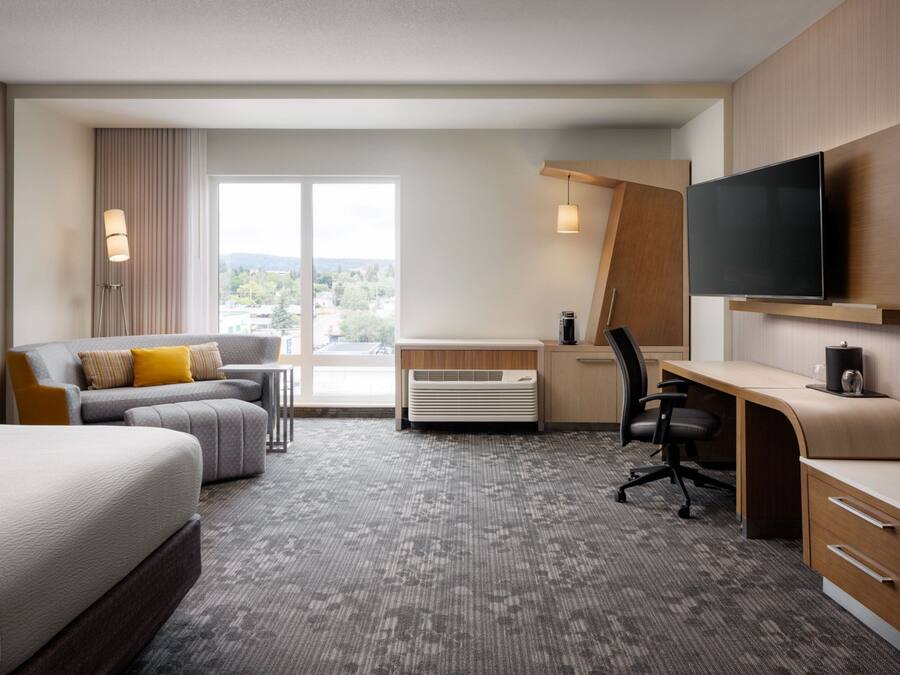 Habitación de hotel con cama, escritorio con silla, sofá, televisor montado en la pared y un amplio ventanal con vistas al exterior.