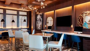 Uma área de bar moderna, com cadeiras brancas e azuis, uma mesa comprida e uma televisão grande.