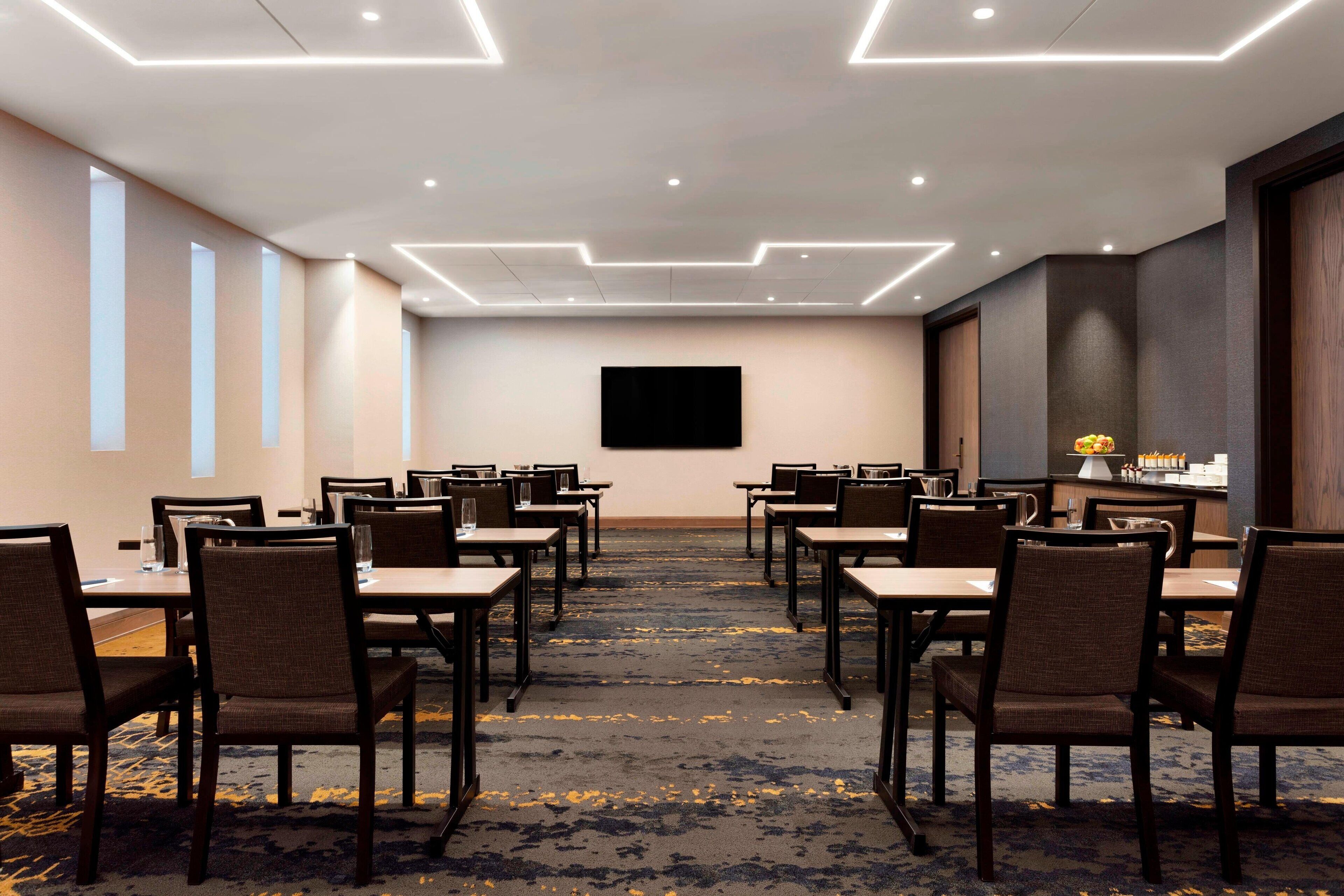 A conference room with tables and chairs arranged for meetings, a large flat-screen TV on the wall, and a carpeted floor.