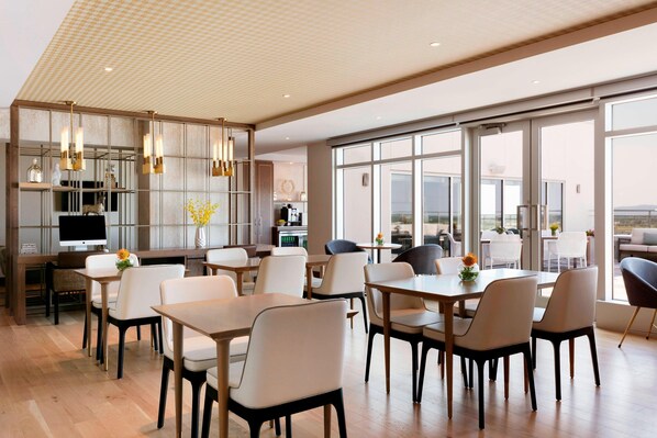 A modern dining area with wooden tables and chairs, a kitchen in the background, and large windows offering a view. - Sheraton Saint-Hyacinthe Hotel (Saint Hyacinthe)