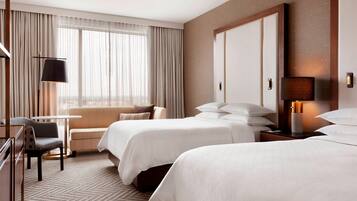 A hotel room with two beds, a desk, a chair, a lamp, and a large window with curtains.