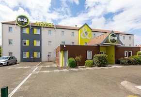 Un hotel B&B con fachada amarilla y marrón, varias ventanas y un estacionamiento.